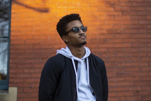 Alejandro stands in sunglasses in front of a brick building